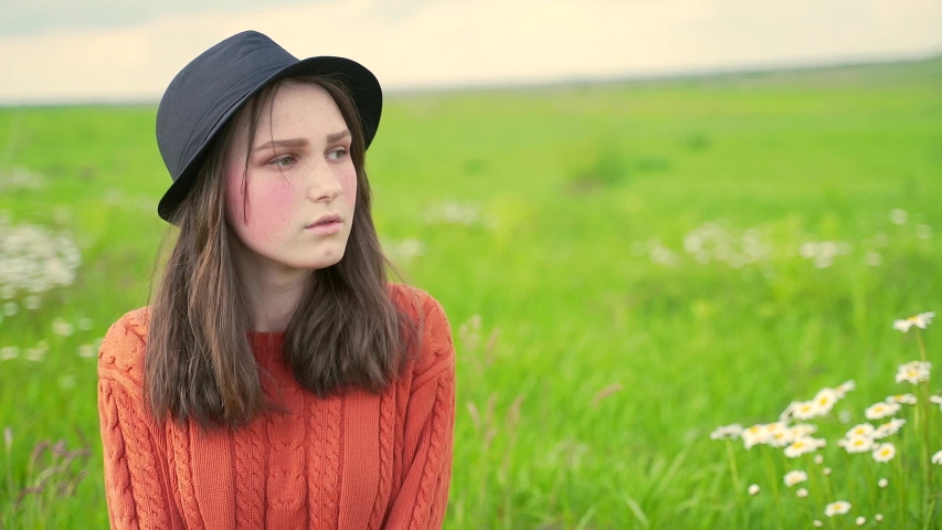 Portrait of a young rural girl in a hat and sweater sitting on the lawn on the grass. Teen girl with freckles and red cheeks in nature with close up. Green eyes and a hole in the chin