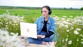 Young girl talking on video call with big black headphones on lawn in park. Stylish teenager smiling and sitting on grass on flowers background in jeans jacket with white computer.