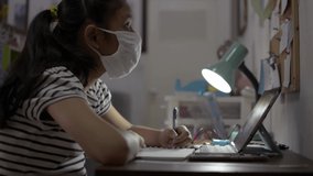 Elementary school girl wearing protective face mask and using digital tablet for lesson online at home. Female teenager writing on a book while studying from home with video call during COVID-19.