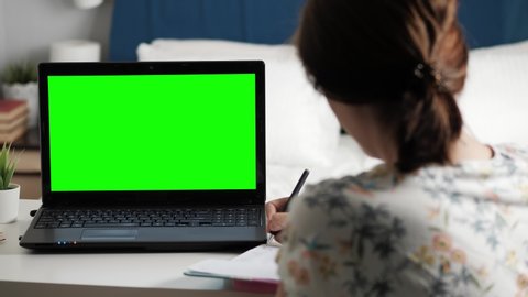 Woman sits at desk in bedroom, she looks at laptop green screen and talks to someone over internet video communications, sometimes taking notes in notebook. Close-up