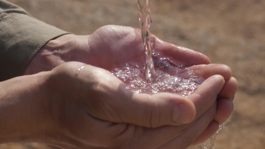 Stream of fresh drinking water pouring into human hands, drought land on the background. dry farmland, lack of water. Clean water splashing on hands of the poor rural man. ecology concept