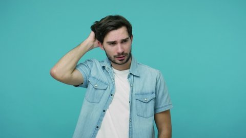 Frustrated confused young bearded guy in jeans shirt scratching head contemplating how to solve problem, thinking intensely with puzzled look, having doubts. studio shot isolated on blue background