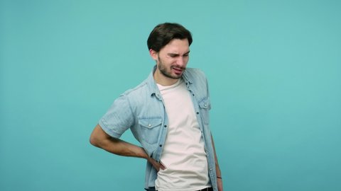 Backache. Tired unhealthy male worker in jeans shirt touching injured sore back, suffering lower lumbar discomfort, muscle pain of overwork, pinched nerve. studio shot isolated on blue background