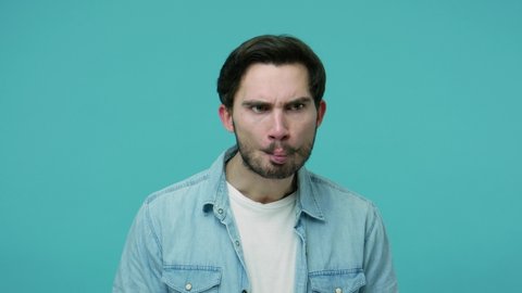 Comical weird bearded guy in jeans shirt watching around making fish face with pout lips, frowning and looking with humorous wary suspicious expression. indoor studio shot isolated on blue background