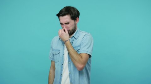 Displeased bearded guy in jeans shirt pinching his nose holding breath, expressing disgust for unpleasant smell, bad breath, avoiding stinky odor. indoor studio shot isolated on blue background