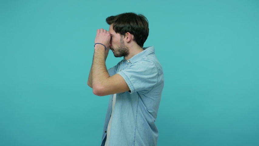 Nosy curious guy in jeans shirt looking through binocular gesture, seeking something in distance, watching far away with amazement and shock, exploring way. studio shot isolated on blue background Royalty-Free Stock Footage #1054232228