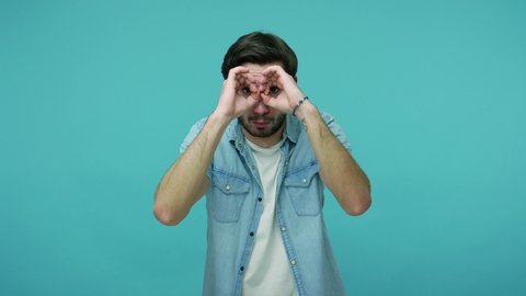 Nosy curious guy in jeans shirt looking through binocular gesture, seeking something in distance, watching far away with amazement and shock, exploring way. studio shot isolated on blue background