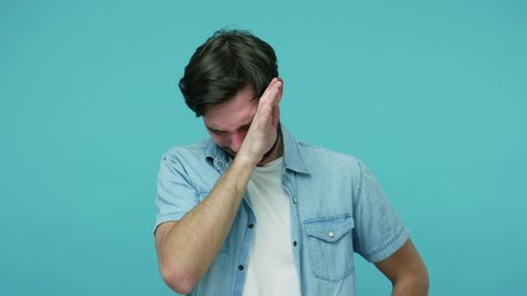 Miserable depressed dramatic bearded guy in jeans shirt crying and wiping away tears, experiencing loss defeat, feeling lonely and desperate in his grief. studio shot isolated on blue background