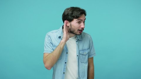 Deafness and hearing problems. Attentive bearded guy in jeans shirt keeping hand near ear and listening carefully to gossip, conversation, secret news. indoor studio shot isolated on blue background