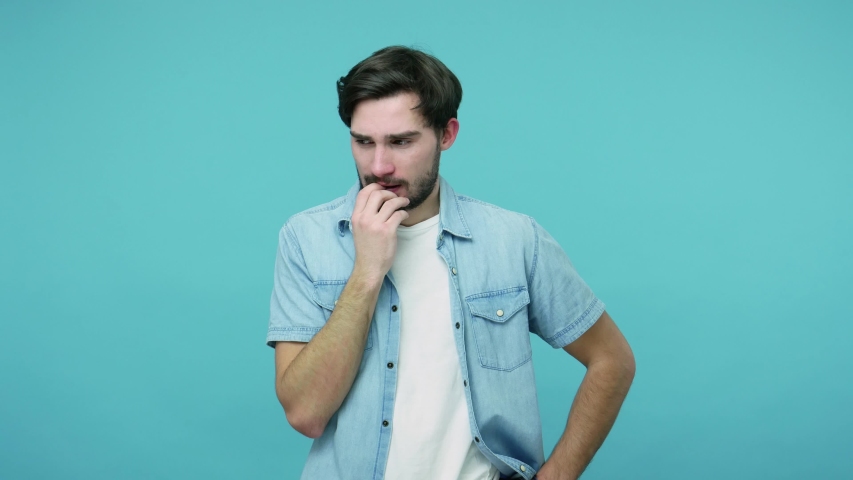 Worried confused guy in jeans shirt rubbing his bristle and thinking over difficult question, expressing doubts, hesitating about difficult choice. indoor studio shot isolated on blue background | Shutterstock HD Video #1054232333