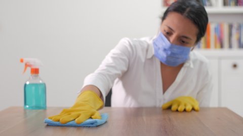 A Hispanic woman wearing a white shirt, a face mask and yellow gloves is cleaning a table with a blue cloth
