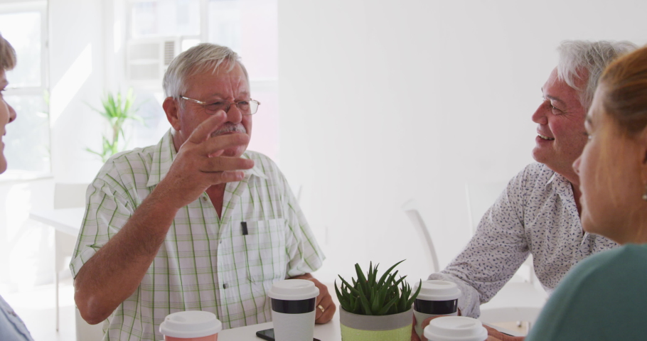 A group of happy senior male and female friends sitting at a table together socialising before their ballroom dancing class, talking and drinking tea and coffee in slow motion