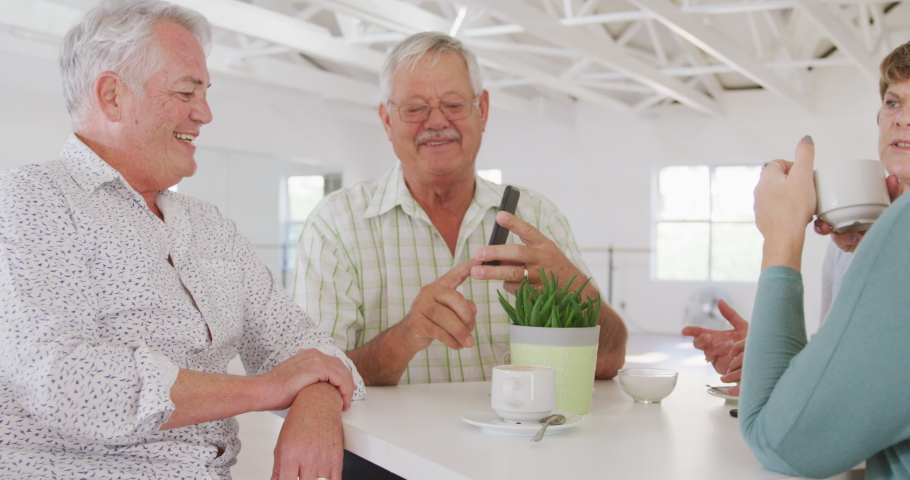 A group of happy senior male and female friends sitting at a table together socialising before their ballroom dancing class, talking and drinking tea and coffee, using a smartphone in slow motion