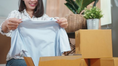 Asian woman is unpacking cardboard box and look inside the box and removing multiple colorful t-shirts from a cardboard box in her new home or apartment. Close up of Female hand unpacking box. 