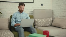 Attractive happy man using laptop sitting on couch in modern living room at home internet business
