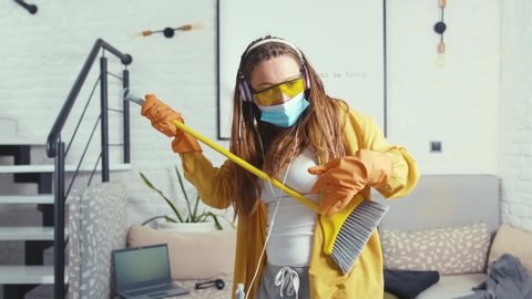 Close up young woman with mask and glasses singing and dancing with mop during clean-up enjoying music in headphones housewife fun beautiful housework cleaning slow motion