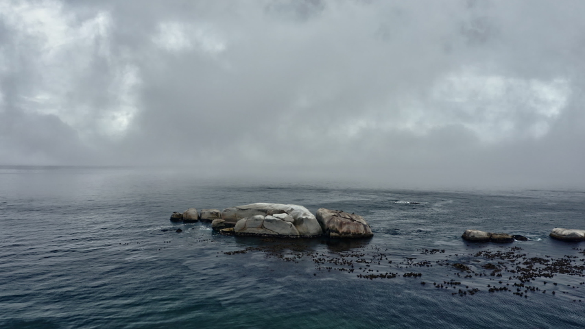 Orbiting shot of the deep blue ocean waters with a huge rock in the middle, thick fog covering part of the frame and a view of the lush mountains in the background.