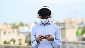 Young Asian / Indian man playing mobile game and listening music on headphone while wearing surgical mask for safety, person protecting himself from corona virus or covid-19 by wearing face mask.