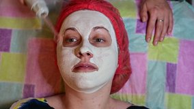 Video of the face of a female patient in a spa salon. A beautician in a dressing gown on a couch makes a facial massage with a mask.