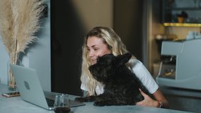 Happy smiling blonde woman waves her hand and makes a video call with friends on the computer. Female owner hugging adorable puppy dog and chatting at laptop camera with parents from home.
