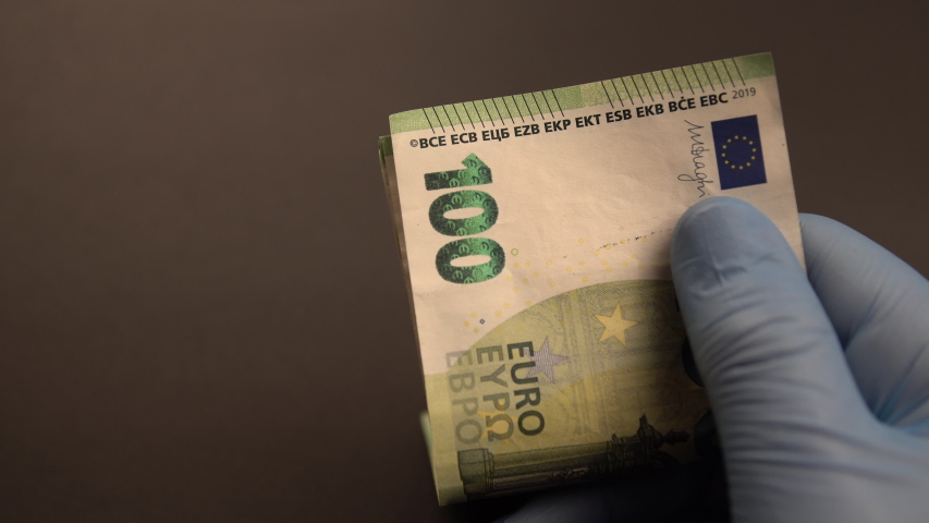 Money exchange during Covid-19 times: Hands with blue protective medical gloves are counting European 100 (hundred) EURO money banknotes and leave them on black background. Royalty-Free Stock Footage #1054259678