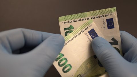 Money exchange during Covid-19 times: Hands with blue protective medical gloves are counting European 100 (hundred) EURO money banknotes and leave them on black background.