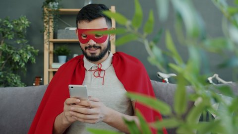 Successful guy in super hero costume is using smartphone smiling sitting on couch in apartment and communicating online. People and devices concept.