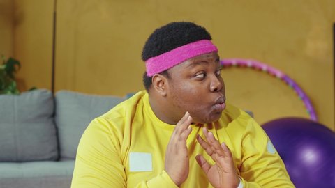 Cunning fat african american man practicing yoga meditation with personal trainer and secretly eating a cookie. Funny scene. Fitness parody. Humor concept.