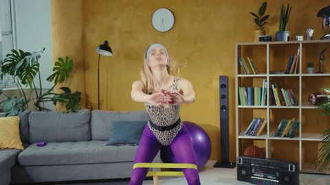 Attractive fit retro sportswoman doing exercises on glutes making squats with elastic resistance bands in the living room. Concept of sport, activity, fitness.