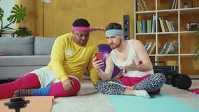 Friendly couple of handsome unfit young men wearing yoga sportswear on mats using smartphone browsing social media during fun fake fitness workout.
