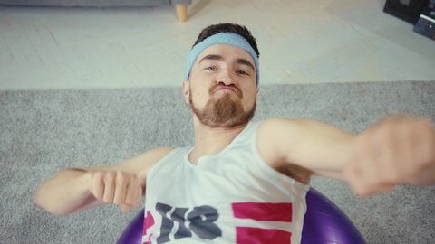 Close-up of funny fitness freak recording comical sports vlog, lifting imaginary barbell up, flexing arms and exercising on fitball. Concept of humor in sports.