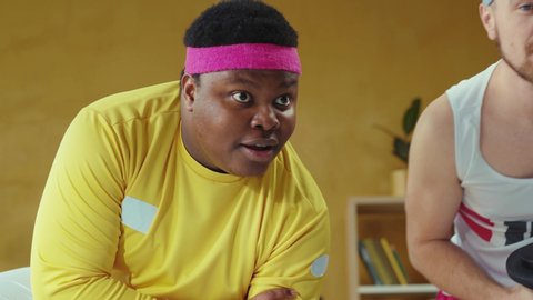 Overweight young african man wearing retro clothing, lifting dumbbells working on muscles, training home workout with friend using online references.