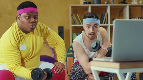 Motivated couple of multi-race unfit guys watching online classes on laptop. Beginner team of retro style men friends going in for sports trying working out with dumbbells.