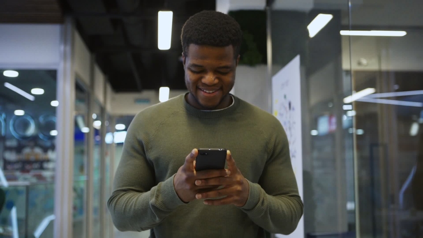 Medium waist up shot of smiling young Black man walking down office corridor in slow motion and enjoying text messaging on cell phone Royalty-Free Stock Footage #1054272011