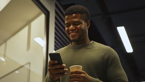 From below medium shot of smiling young Black man with disposable coffee cup walking down office corridor in slow motion and enjoying text messaging on cell phone - Powered by Shutterstock - Get 15% off with code: PIKWIZARD15