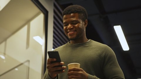From below medium shot of smiling young Black man with disposable coffee cup walking down office corridor in slow motion and enjoying text messaging on cell phone