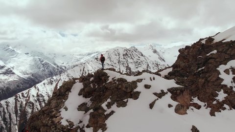 A male traveller with a backpack stands on top of a cliff against the backdrop of snow-capped mountain peaks and low clouds. Aerial view. Flying the camera around a man in a storon cliff