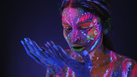 Portrait of girl with a UV pattern on her face, she slowly moves her hands and looks with black eyes at the camera. Graceful model posing with UV pattern on body. Body art glowing in ultraviolet light