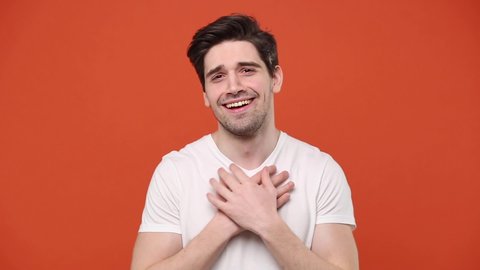 Young cute guy 20s 30s years old in white t-shirt isolated on orange background studio. People emotions lifestyle concept. Looking at camera charming smile ask who me oh it so sweet put hands on chest
