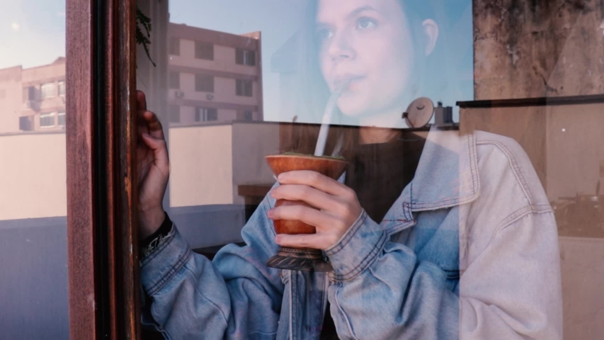 Reflective, Pretty Woman Drinking A Chimarrão Standing By Window At Home