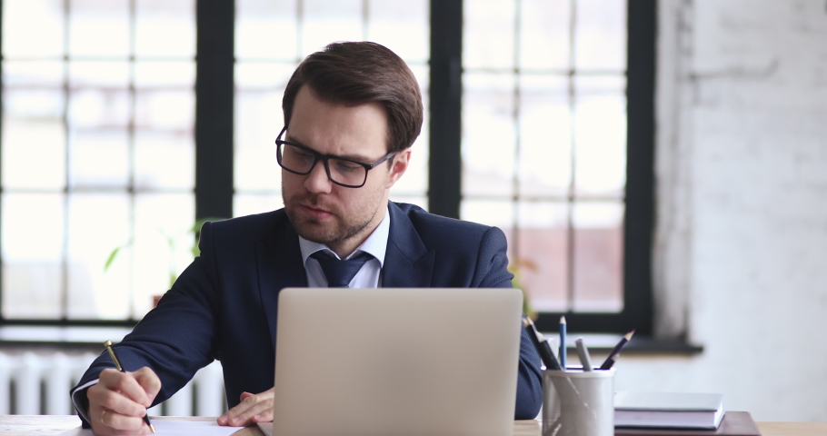 Thoughtful 30s male executive manager in glasses looking away, developing corporate growth strategy. Pensive businessman in suit solving problems, writing notes in paper planner and online apps. Royalty-Free Stock Footage #1054280738