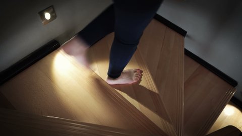 A barefoot female walking down over a wooden stairs lit by led lights at night in luxury home