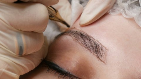 A special needle tattoo machine makes permanent makeup correction of a young woman's eyebrows. A pigment of dark paint is injected under the skin. Microblading, powder spraying close up.
