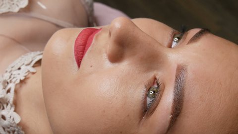 Portrait of a beautiful young woman with permanent make up microblading with eyebrow tattoo and lips with red lipstick lies on a couch in a beauty salon after the procedure.