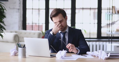 Stressed overwhelmed young businessman in suit working on computer, suffering from eyes fatigue or having painful feelings. Exhausted executive manager overworked in office, burnout concept.