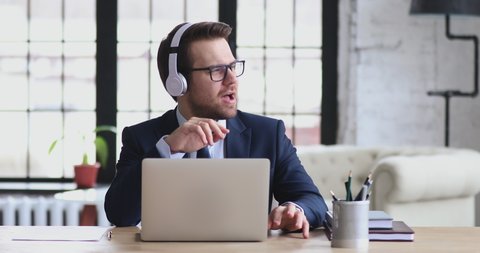 Funny young company representative manager sitting at workplace, listening to favorite music in wireless headphones. Happy relaxed 30s businessman enjoying break time, singing song at office.