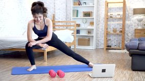 20s young Asian woman in sportswear doing stretching exercises while watching fitness training class on computer laptop online. Healthy girl exercising in living room with sofa couch and bed.