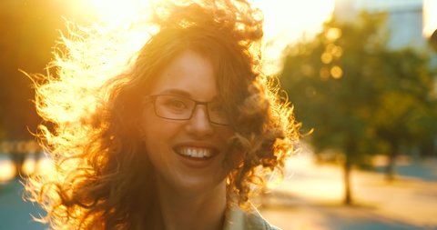Girl walks at sunset light, sun is shining, woman turns back with smile looking at camera. She is free and happy. Curly girl in beautiful sun rays, wind blows her hair. Slow motion