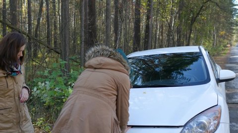 car broke down in the forest, two women open the hood and look for damage. Woman calling road assistance