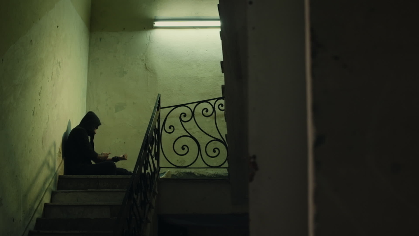 Social issues and drugs, young man injecting heroin with syringe and sitting on stairs of abandoned house. Junkie with addiction, substance abuse, drug addict